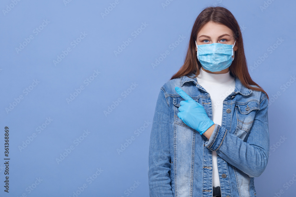 Young attractive dark haired fashionable girl wearing medical mask and latex gloves points aside with her index finger, looking at camera, poses against blue wall. Young attractive dark haired fashionable girl wearing medical mask and latex gloves points aside with her index finger, looking at camera, poses against blue wall.