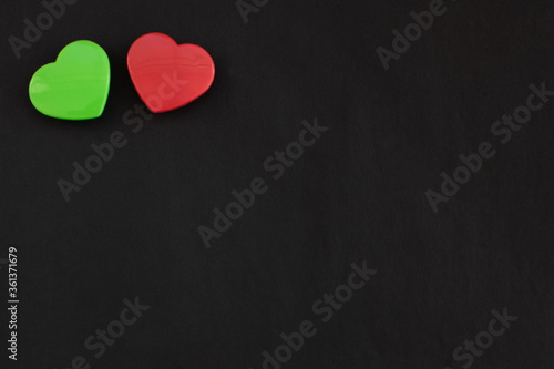 Two green and red color hearts lying close together isolated at the top left corner of black paper background. Flat lay with empty space for text.