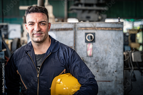 Man in blue jumpsuit holding yellow hard hat in the in the factory. Confident mechanic ready for work.	

