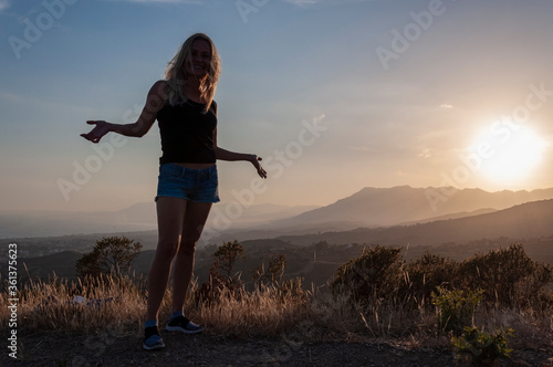 A girl in a black T-shirt, jeans shorts and hats, with her hands spread apart, stands on top of a hill against the backdrop of sunset and mountains. © Andrei
