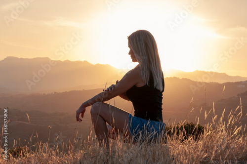 Side view of a blonde girl in a T-shirt, shorts and sneakers squatting among ears of grass against the backdrop of the setting sun and mountain peaks.