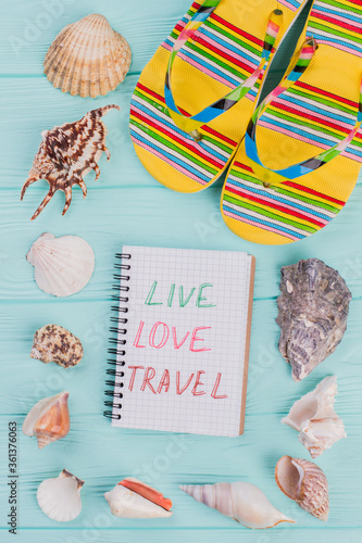 Different sea shells and yellow sandals in the corner on turquoise background. Love live travel written on notepad.