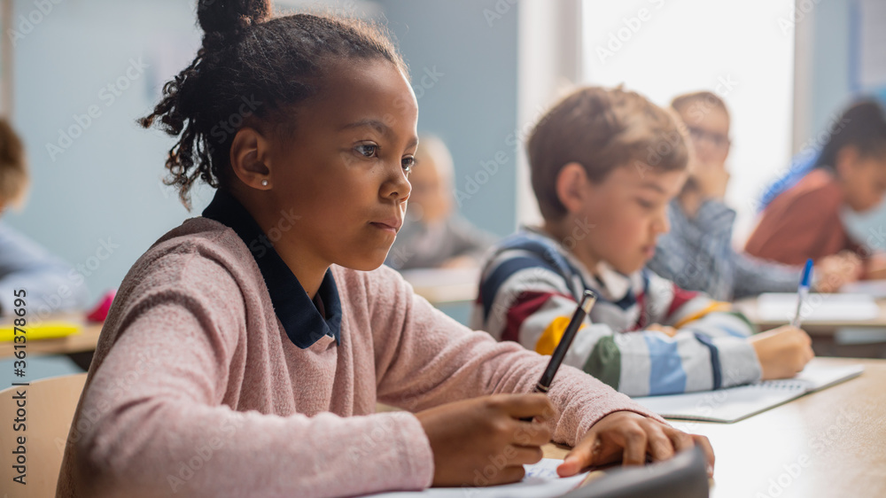In Elementary School Classroom Brilliant Black Girl Writes in Exercise Notebook, Taking Test and Writing Exam. Junior Classroom with Diverse Group of Children Working Diligently and Learning New Stuff In Elementary School Classroom Brilliant Black Girl Writes in Exercise Notebook, Taking Test and Writing Exam. Junior Classroom with Diverse Group of Children Working Diligently and Learning New Stuff