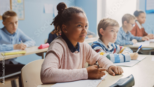 In Elementary School Classroom Brilliant Black Girl Writes in Exercise Notebook, Taking Test and Writing Exam. Junior Classroom with Diverse Group of Children Working Diligently and Learning New Stuff photo