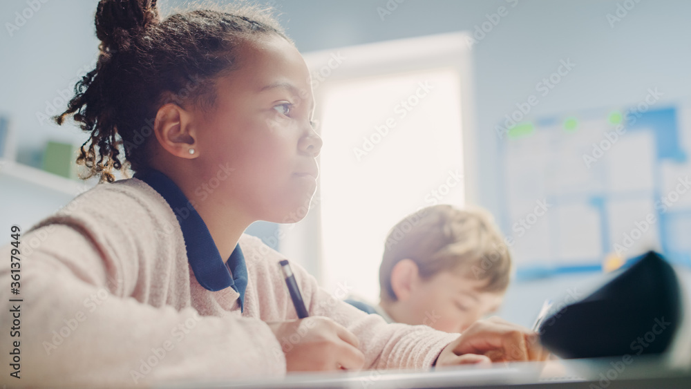 In Elementary School Classroom Black Girl Writes in Exercise Notebook, Taking Test. Junior Classroom with Diverse Group of Bright Children Working Diligently, Learning. Low Angle Side View Portrait In Elementary School Classroom Black Girl Writes in Exercise Notebook, Taking Test. Junior Classroom with Diverse Group of Bright Children Working Diligently, Learning. Low Angle Side View Portrait