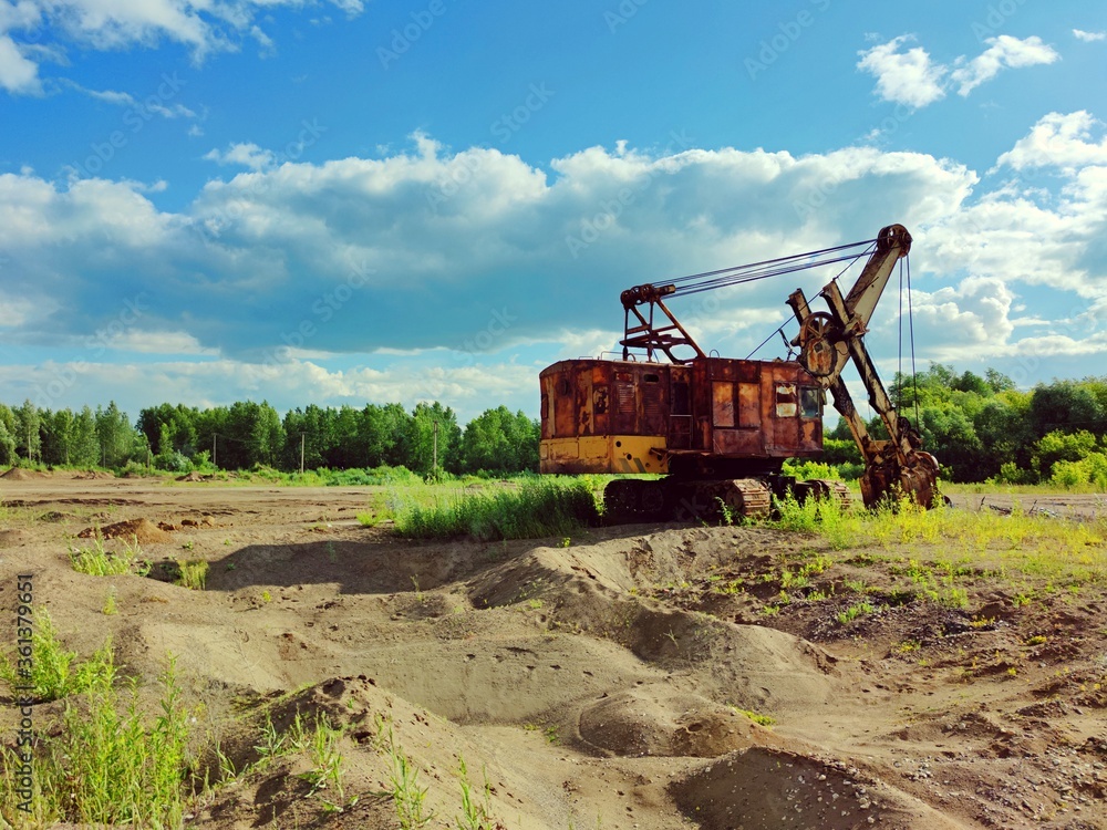 Obraz premium old rusty excavator on a sand quarry on a background of green forest and blue sky on a sunny day