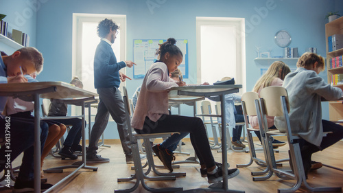 In Elementary School Class: Enthusiastic Teacher walks between Rows of Diverse Children, Explains Lesson. Group of Smart Multiethnic Kids Learning New Stuff. Side View Shot