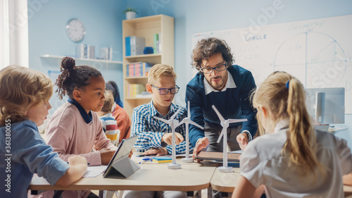 Elementary School Classroom: Enthusiastic Teacher Holding Tablet Computer Explains to a Brilliant Young Children How Wind Turbines Work. Kids Learning about Eco-Friendly Forms of Renewable Energy