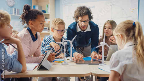 Fotografie, Obraz Elementary School Classroom: Enthusiastic Teacher Holding Tablet Computer Explains Lesson to Brilliant Young Children