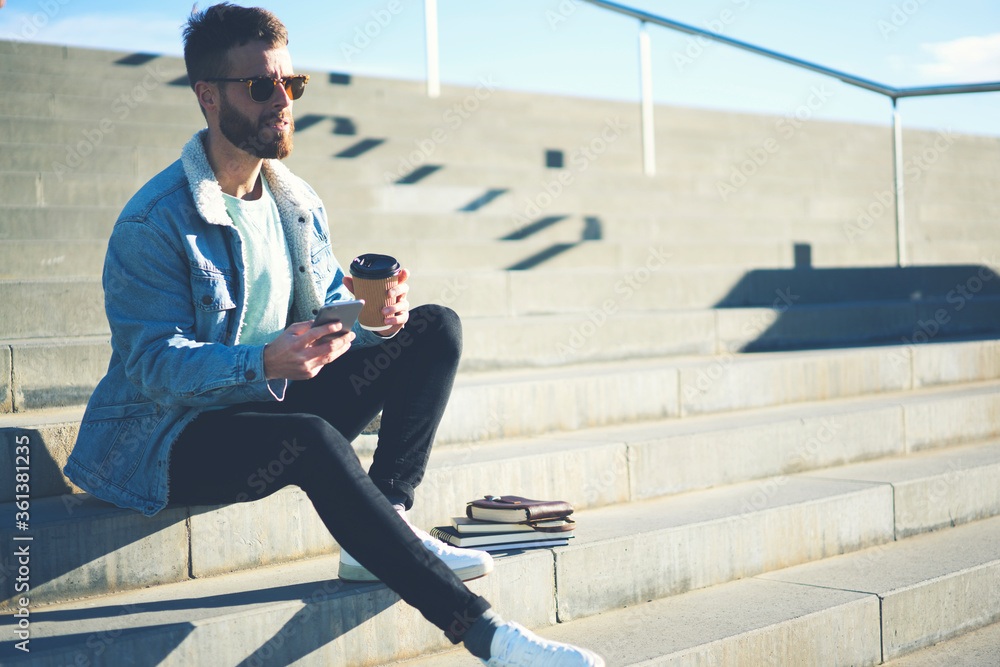 Young handsome international student in casual street wear outfit enjoying coffee on break in university spending time outdoors chatting with classmates via smartphone and 5G wireless connection Young handsome international student in casual street wear outfit enjoying coffee on break in university spending time outdoors chatting with classmates via smartphone and 5G wireless connection