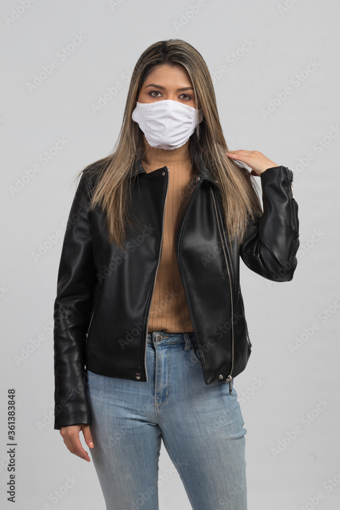 latin woman protecting her face with white mask, using black leather jacket and jeans, studio with gray background latin woman protecting her face with white mask, using black leather jacket and jeans, studio with gray background