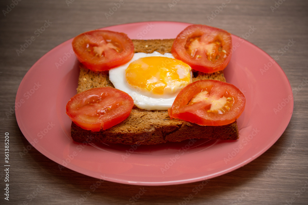 Toast bread with fried egg and sliced tomatoes on pink dish on wooden table.