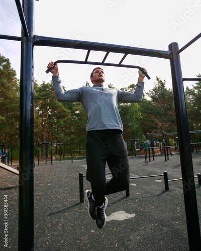Muscular sports trainer on street workout exercising. Fit and athletic man working out in an outdoor gym. Pull ups on horizontal bar.