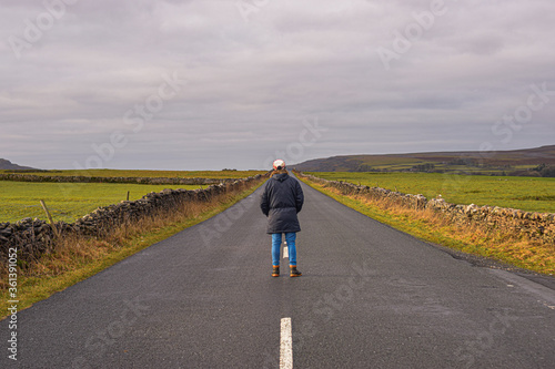 Photo of a man with a winter coat standing in the middle of the road surrounded by nature in the UK