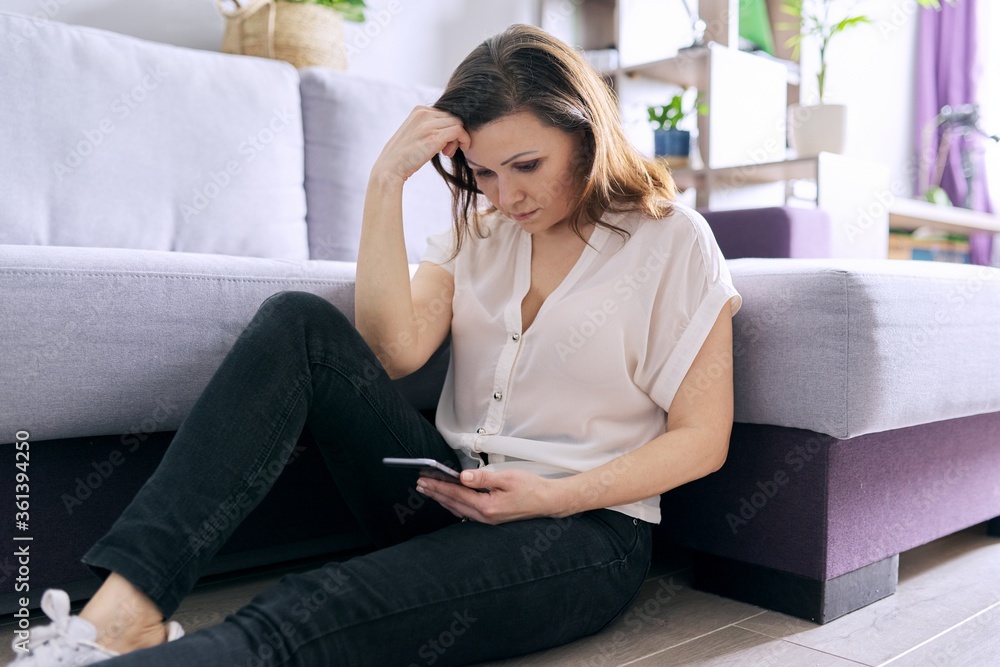 Mature sad woman reading smartphone sitting at home on the floor Mature sad woman reading smartphone sitting at home on the floor