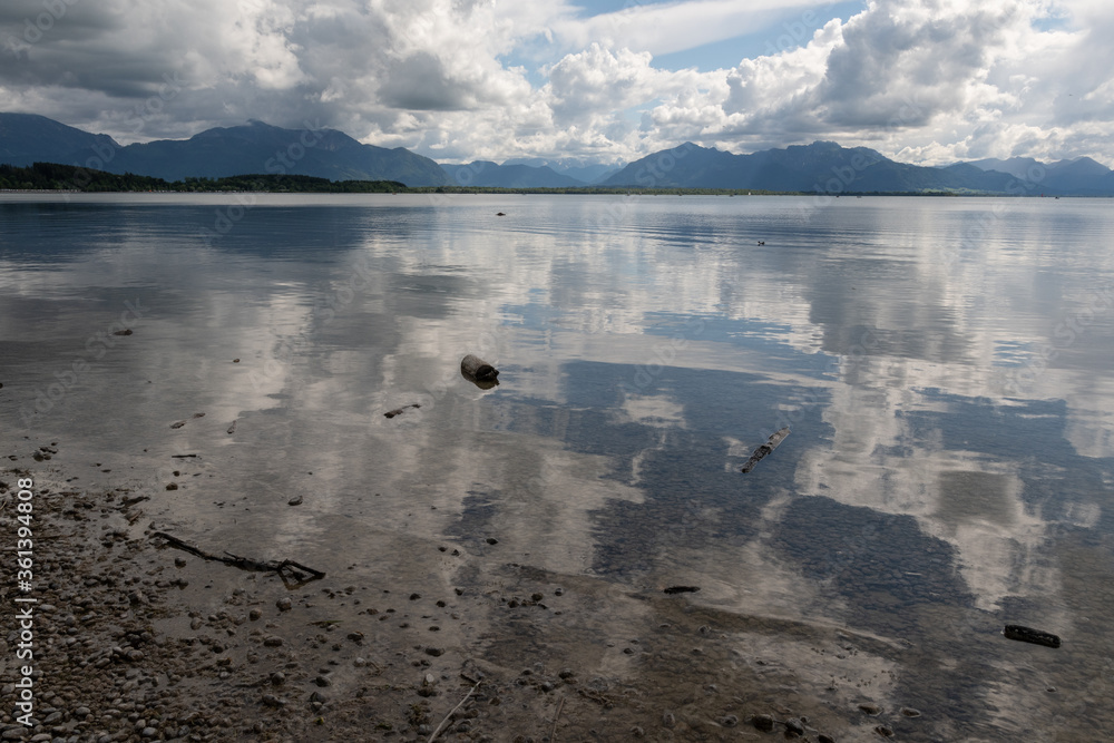 Fototapeta premium Spiegelungen Chiemsee mit Bergen , Himmel und Wolken und schwimmenden Baumstämmen