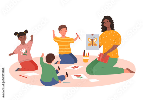 Dedicated African-American female teacher showing to the kids a hand-drawn butterfly, while teaching them drawing and colouring on paper in a kindergarten with children of various nationalities