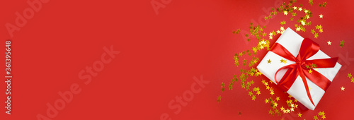 Gift box, golden stars confetti  on red background. Greeting composition for Christmas, New year, Valentines day or birthday. Minimalist concept. Banner with copyspace.