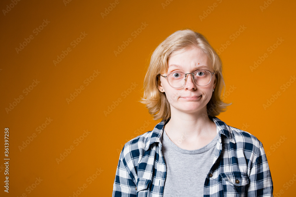 Blonde girl in a plaid shirt with glasses and hard looks into the camera on an orange background.