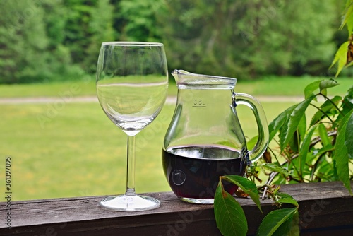 An empty wine glass and a jug of red wine on a railing. Terrace in the middle of a forest. Wooden railing overgrown with green leaves.