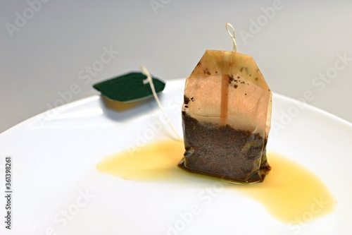 One bag of black tea on a white plate. Used tea bag on a plate. Tea bag on a plate with a puddle of tea. Gray and white background.