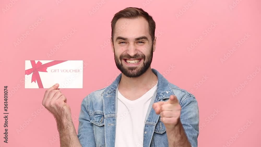 Bearded cute young man guy 20s in denim jacket white t-shirt isolated on pastel pink background studio. People lifestyle holiday concept. Look camera with charming smile hold in hand gift certificate Bearded cute young man guy 20s in denim jacket white t-shirt isolated on pastel pink background studio. People lifestyle holiday concept. Look camera with charming smile hold in hand gift certificate