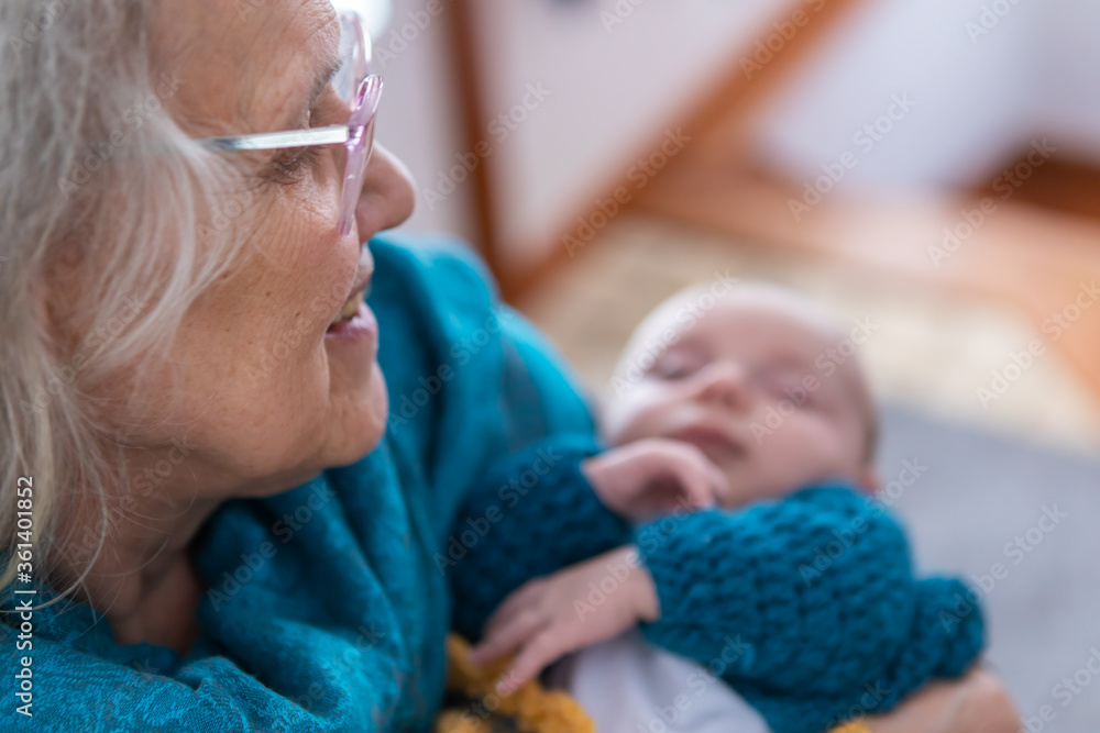 Selective focus portrait of senior woman in pink glasses gazing at her grandchild, out of focus we see a small child swaddled in her arms. Selective focus portrait of senior woman in pink glasses gazing at her grandchild, out of focus we see a small child swaddled in her arms.