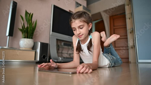 A girl in a white T-shirt and jeans lies on the floor of the room and plays on the tablet. The girl raises and shakes her legs.