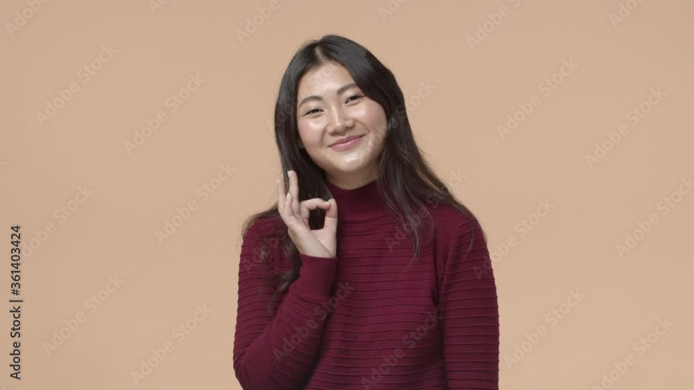 Okay deal. Silly attractive brunette korean girl over beige background, showing OK sign, agree or approve something, nod positive reply, satisfied with plan, recommend or being pleased Okay deal. Silly attractive brunette korean girl over beige background, showing OK sign, agree or approve something, nod positive reply, satisfied with plan, recommend or being pleased