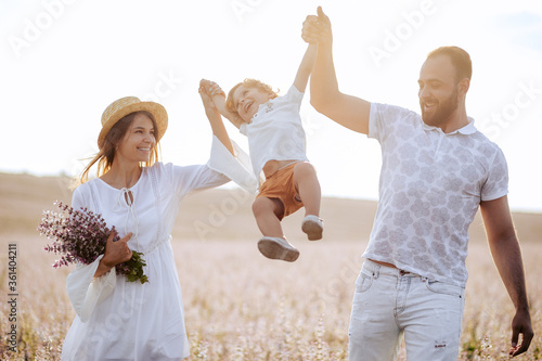 Happy family portrait outdoor at the sunset time. People having fun on the field. Concept of friendly family and of summer vacation. Parents and son spending good time. White dress. Straw hat. Kissing