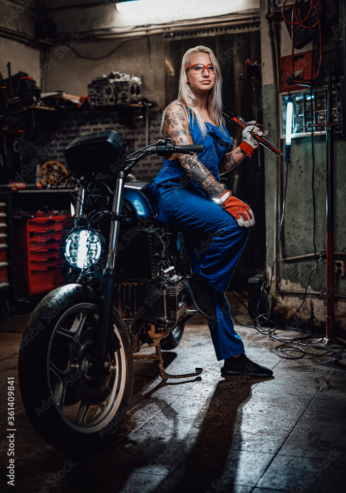Tattooed female mechanic in work overalls hold a big wrench and posing for a camera while leaning on her naked bike in garage or workshop