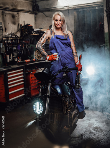 Tattooed hipster girl in work overalls hold a big wrench while standing on naked bike in garage or workshop, smiling and looking on camera