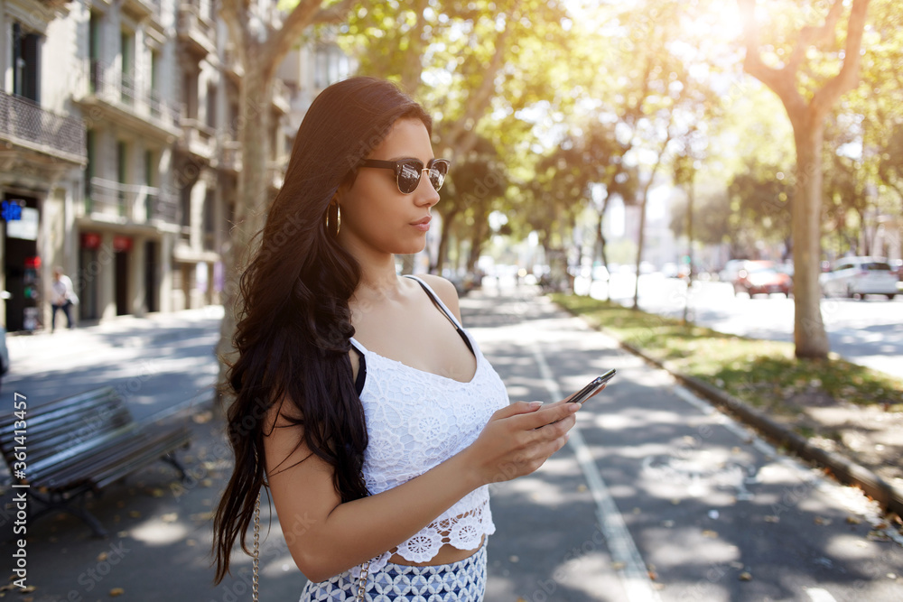 Gorgeous Hispanic woman chatting on mobile phone while standing in the street in summer day, pretty female with long brunette hair reading news on cell telephone while waiting for boyfriend outdoors