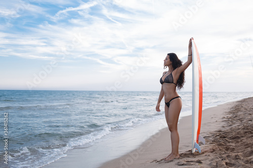 Full length portrait of sexy female surfer standing on the background of a tropical beach landscape, gorgeous young model with beautiful figure waiting for a wave to surf on it and get some pleasure