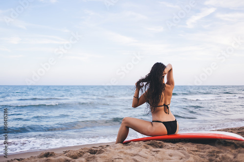 Back view of beautiful girl sitting on a surfboard in sexy bikini and adjusts his wet hair after swimming in summer day, attractive female model posing in swimwear for the magazine of women's clothing