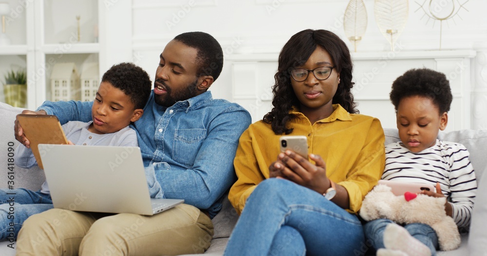 Happy african american family sitting on sofa together with device at home. Mother and father with two little childrens using laptop and smartphone. Happy african american family sitting on sofa together with device at home. Mother and father with two little childrens using laptop and smartphone.