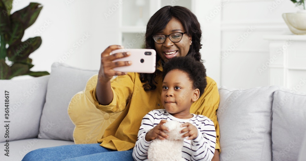 Happy smiling african american mother and cute little daughter taking selfie on smartphone sitting on sofa in living room at home. Happy smiling african american mother and cute little daughter taking selfie on smartphone sitting on sofa in living room at home.