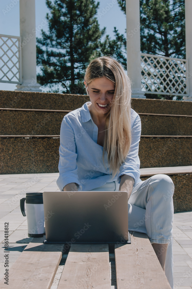 Business lady talks on the phone. A young business woman is working with a laptop in a blue shirt on the street in a Park under the sun! Business concept Business lady talks on the phone. A young business woman is working with a laptop in a blue shirt on the street in a Park under the sun! Business concept