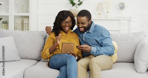 Happy African American couple husband and wife sitting on sofa using tablet and credit card for online shopping at home. © VAKSMANV