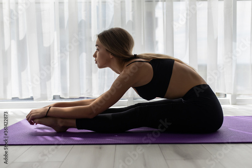 Side view of a woman exercising and stretching her body doing flexibility exercises and bending her back, sitting on a yoga mat indoors.