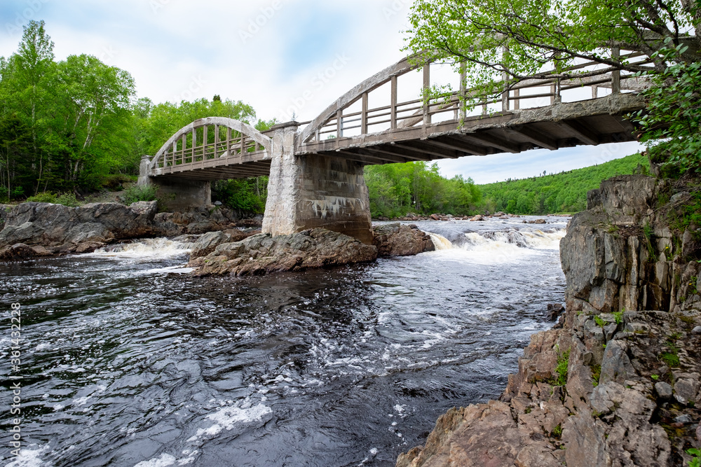 Fototapeta premium Vintage concrete arch bridge with two spans. There's trees on both sides of the bridge with large rock walls of slate. The river is a salmon river with multiple runs. It's a sunny summer's day.