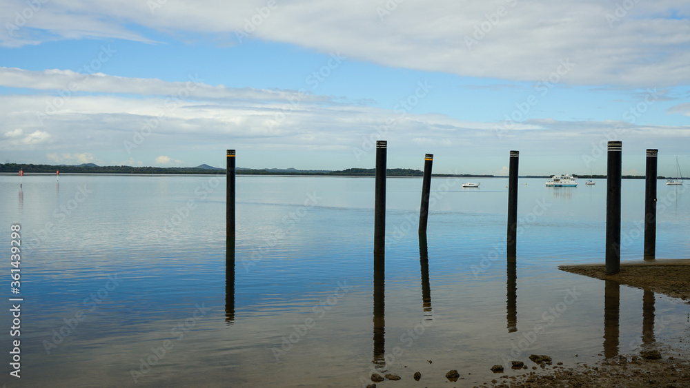 Beautiful calm seascape with blue sky, clouds, and poles reflected in the water. Victoria Point, Moreton Bay, Queensland, Australia.
