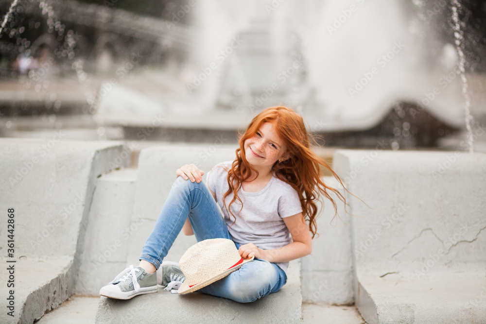 Cute cheerful red-haired child girl with a straw hat sitting by the fountain in the summer outdoors. Summertime in the city