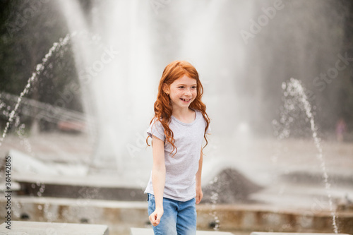 Cute red-haired child girl has fun at the fountain in the summer outdoors. Summertime in the city