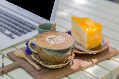 Close up cup of coffee with cake and laptop on table.