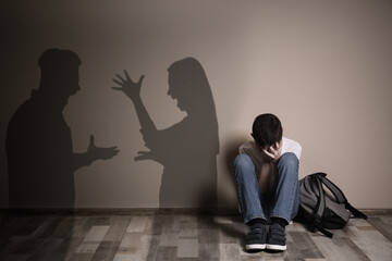 Upset boy with backpack sitting on floor and silhouettes of arguing parents