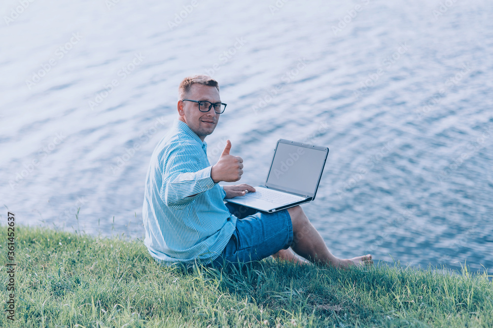A man in glasses and a shirt working on a laptop in the fresh air, against the background of a river.