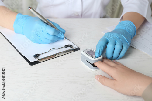 Фотография Doctor examining patient with fingertip pulse oximeter at white wooden table, cl