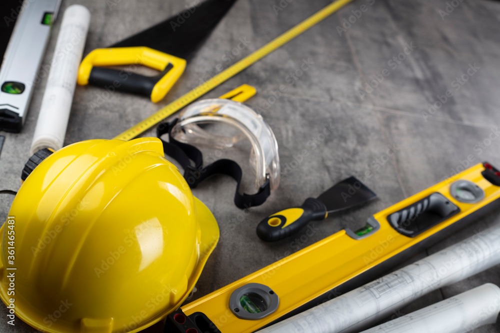 Contractor concept. Tool kit of the contractor: yellow hardhat, libella, hand saw. Plans and notebook on the gray tiles background.