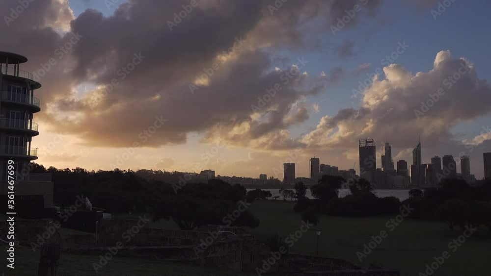 Storm clouds swirl above a windy Perth City just before dusk. In foreground sunlight has almost faded over green park wth many trees. Swan River lies between city and park Storm clouds swirl above a windy Perth City just before dusk. In foreground sunlight has almost faded over green park wth many trees. Swan River lies between city and park